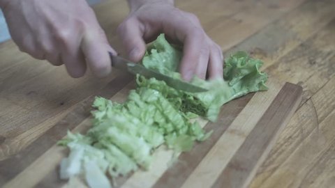 Hands of a man chop green salad, on a wooden table, with a small knife.
