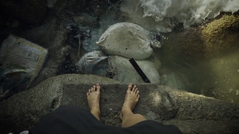 High Angle Shot of Person's Legs Standing on a Rock Near the Sea with Garbage and Trash Floating near Shore. Shot on RED Epic 4K UHD Camera.