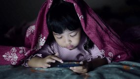Asian kid girl playing game on the tablet or phone under blanket at the night time.Digital Tablet Device Late After Bedtime concept.