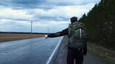 A tourist hitchhiker walks along the road with a backpack on his shoulders, lifting his thumb up. Traveler is trying to stop the car. The weather is cloudy and rainy.