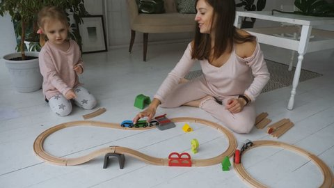 Little adorable girl and her mother is playing with wooden toy railroad while sitting on the floor in white living room