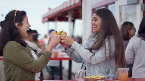 Best Friends Cheers With Their Burgers, Then Really Enjoy Taking First Bite, On Santa Monic Pier (Shot On Red Scarlet-W Dragon In 4K, Slow Motion)