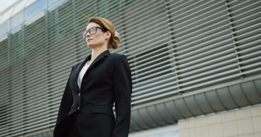 Portrait shot of the Caucasian attractive business lady posing to the camera and crossing her hands while standing on the big office building background. Outdoors