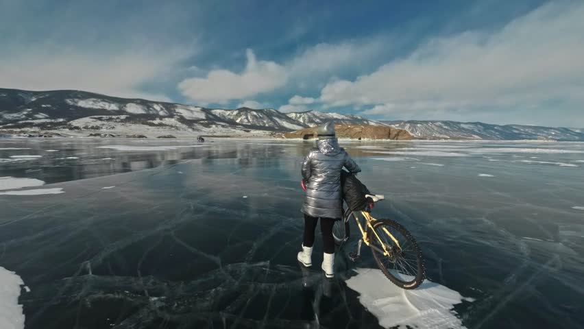 Woman is walking beside bicycle on the ice. The girl is dressed in a silvery down jacket, backpack and helmet. Ice of the frozen Lake Baikal. The tires on the bicycle are covered with special spikes