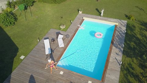 AERIAL: Young Caucasian woman sunbathing tries to break the stalking drone filming her while she relaxes by the pool in her backyard. Buzzing remote controlled plane annoys girl who tries to crash it.