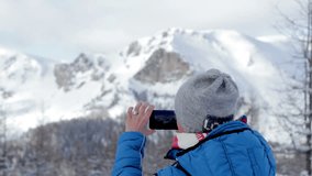 Young woman uses a smartphone to take a picture of beautiful view in the snowy mountains, rear view
