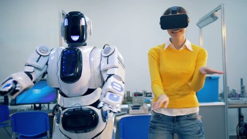 A young female and a cyborg are moving their hands and hips in synch while dancing
