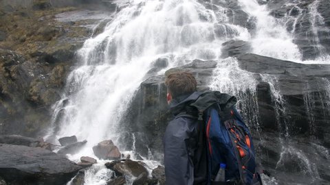 4K Young handsome traveller standing in front of beautiful waterfall in Scottish Highlands. Man hiking backpacker through Scotland UK. Travel and adventure in Europe. 