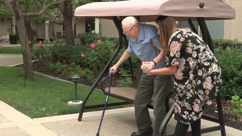 A friendly female caretaker helps an old man with mobility issues out of an outdoor swing