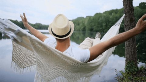 Young man by the lake hanging on hammock relaxing in the morning and stretching arms outstretched . People relaxation travel concept.  SLOW MOTION