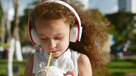 Pretty caucasian little girl curly hair listening to music in white headphones and drinking coconut.