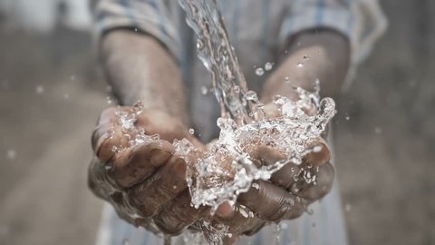 slow motion shot of pure and freshwater water falling on poor man's hands against barren and dry farmland. Clean drinking water splashing on hands of the poor farmer in a drought affected area 