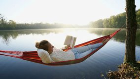 Young woman by the lake hanging on hammock reading book in the morning. People relaxation travel technology concept.  4K