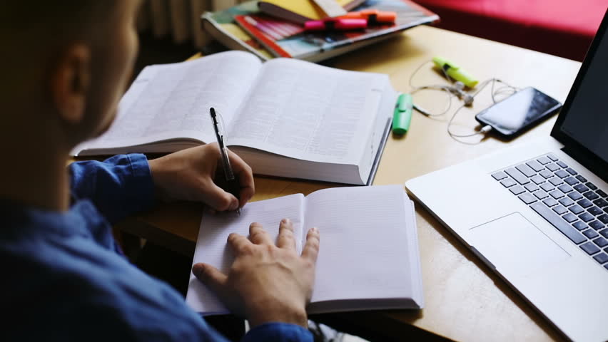 closeup man hand writing something in blank notebook on wooden table with smartphone headphones and laptop on blur background 