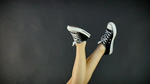 Female legs in classic black and white sneakers. Legs are raised upwards and dangle from side to side. Gray, black background. Classic shoes, retro style. Healthy lifestyle