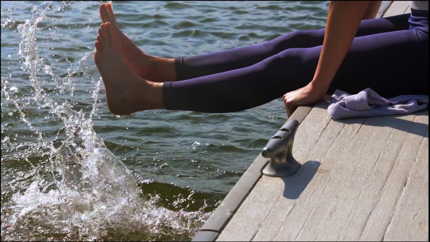 slow motion young woman sitting on a dock and splashing her feet in the lake