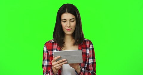 Portrait of the brunette good looking young woman in the red motley shirt scrolling and taping on the tablet device on the chroma key background. Green screen.