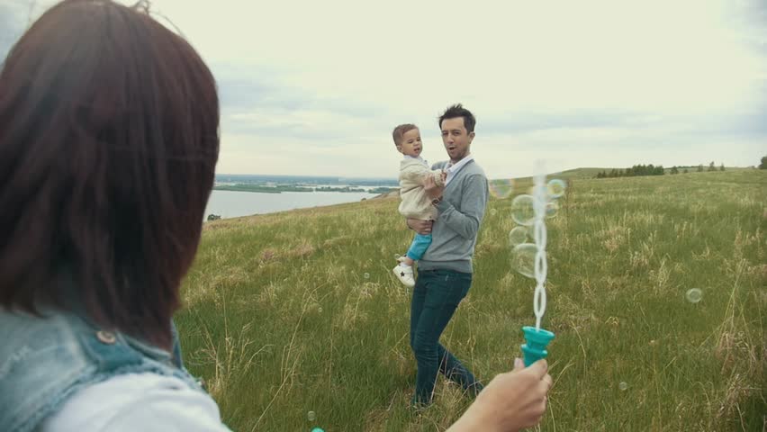 A young family plays with a child on the field, a mother blows bubbles, a dad with a child catches them and laughs