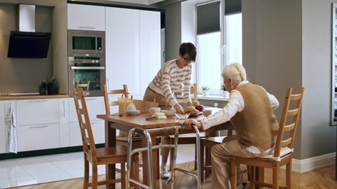 Medium shot of young female caregiver helping infirm elderly lady to sit down at kitchen table, then serving her breakfast