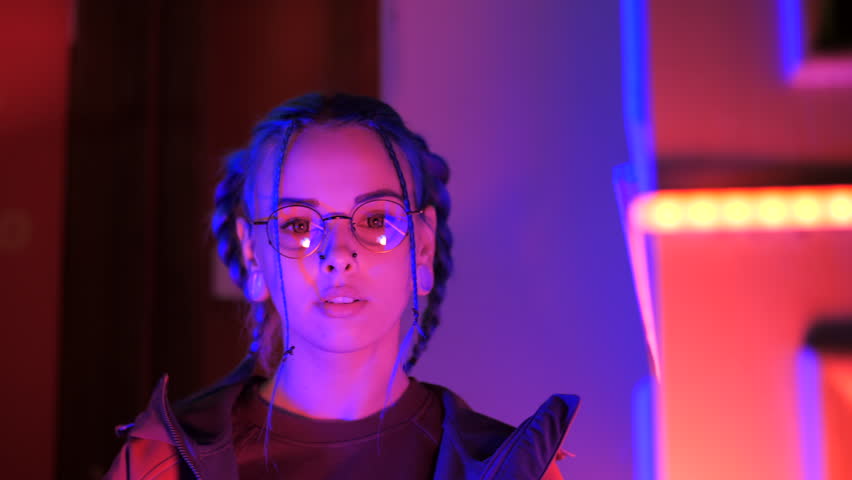 Young pretty girl with unusual hairstyle near glowing neon lights of the city at night. Dyed blue hair in braids. Serious hipster teenager in glasses and beautiful lenses.