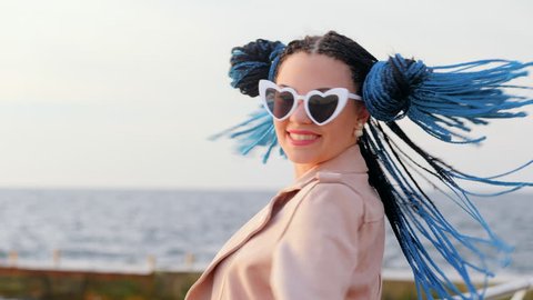 Portrait of young beautiful woman with african kanekalon blue braids posing, smiling to camera on sea beach. Afro hairstyle, dyed hair. Hipster girl in heart shaped glasses.