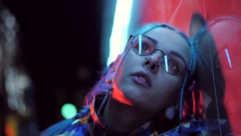 Millennial sad pretty girl with unusual hairstyle near glowing neon of the city at night. Dyed blue hair in braids. Mysterious hipster teenager in glasses. Reflection of light.