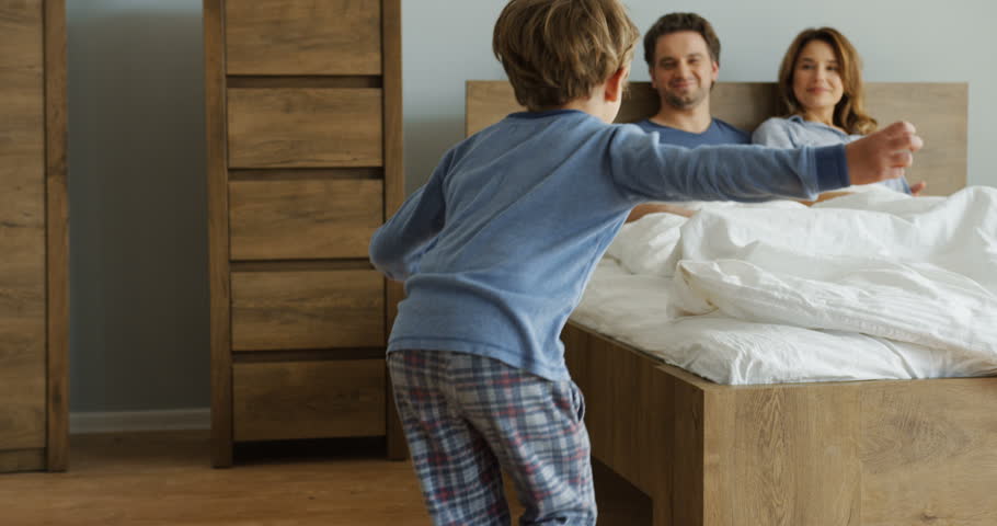 Cheerful little children jumping in the bed to their happy mother and father sitting in the morning in it after waking up. Indoors Royalty-Free Stock Footage #1011486032