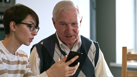 Tilt up of cheerful young woman in glasses showing mobile phone to senior man and teaching him how to use it