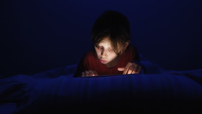Dolly shot caucasian girl lying in bed playing tablet in social internet in dark blue light under blacket. Childhood, communication and technology concept