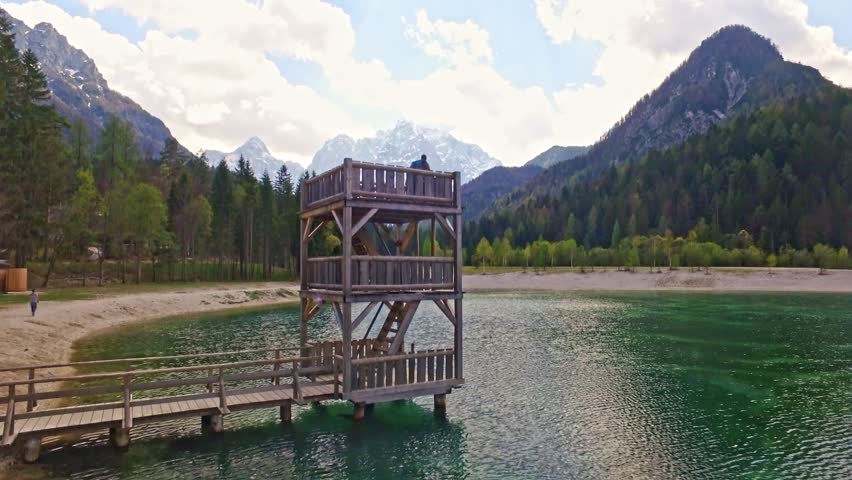 The drone flies vertically, shows a wonderful view of the Slovenian protected natural park with a two story wooden diving board with a young couple standing on top.