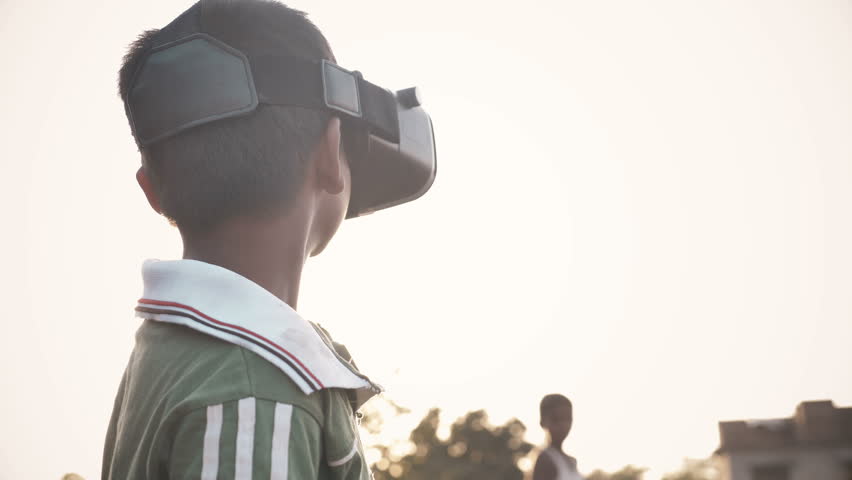 A young kid wearing VR glasses enjoying and looking around in amazement in an outdoor open field against the sun. An excited young boy wearing Virtual Reality glasses in rural India Royalty-Free Stock Footage #1011505298