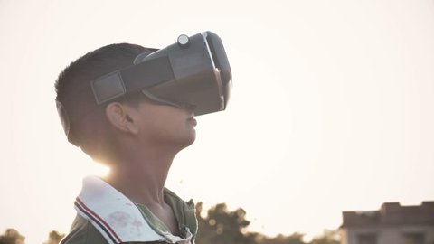 A young kid wearing VR glasses enjoying and looking around in amazement in an outdoor open field against the sun. An excited young boy wearing Virtual Reality glasses in rural India