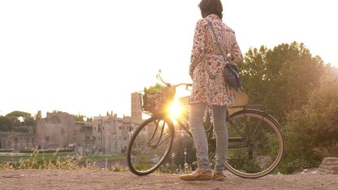 Young woman with bycicle takes pictures of the colosseum in rome at sunset with smartphone. Stylish dress with large hat, flowers and bread in basket.