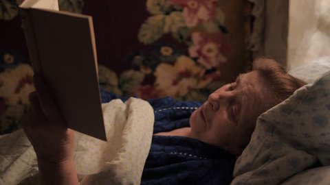 Old elderly woman with wrinkles lying in bed under a blanket and reading a book before sleep, 4K, 3840x2160