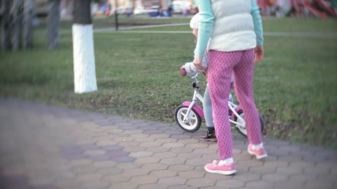 A laughing, smiling mother pushes her daughter forward on a warm spring day, when she teaches her to ride a bike along the city's sidewalk near a green park. 4k