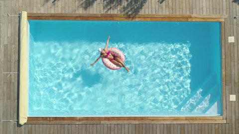AERIAL, TOP DOWN, CINEMAGRAPH: Cheerful young woman sunbathing while relaxing on a cool inflatable doughnut in her backyard pool. Happy girl in pink bikini lying on a funny floatie in amazing pool.