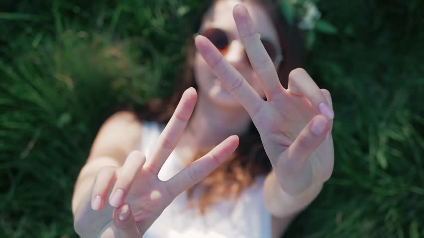 SLOW MOTION: Top view shot of a Caucasian girl in a white floral dress lying in grass and shows a sign of peace