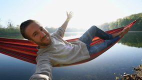 Young man by the lake hanging on hammock relaxing in the morning takes a selfie portrait. People relaxation travel concept.  4K