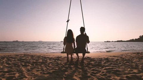 romantic getaway, couple in love sitting together on rope swing at sunset beach, silhouettes of young man and woman on holidays or honeymoon