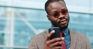 Portrait of young african american man talking using smartphone video chat technology stands next to the office center dressed in a business suit with sunglasses
