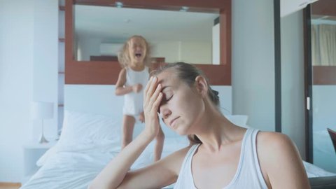 Portrait of upset tired mother with little child girl jumping on bed on the background. A hyperactive difficult kids and despairing parents concept.