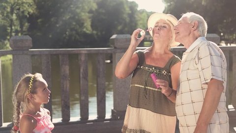 Child and her grandparents outdoors. Cute couple of senior people with bubble blower on bridge, slow motion. Granddaughter and her grandparents spending good time together.