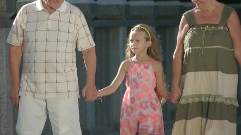 Grandparents walking with their granddaughter. Slow motion child holding hands with her grandparents and walking on bridge, girl looking at her grandmother. Family leisure and relationship.