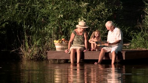 Grandparents and child near river. Elderly people sitting near water with granddaughter. Grandfather tutoring grandchild with book on nature background.