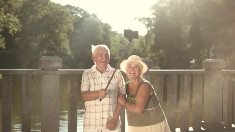 Cute couple of seniors taking a selfie. Elderly man and woman with monopod on nature background in sunny day. Old people and gadget.