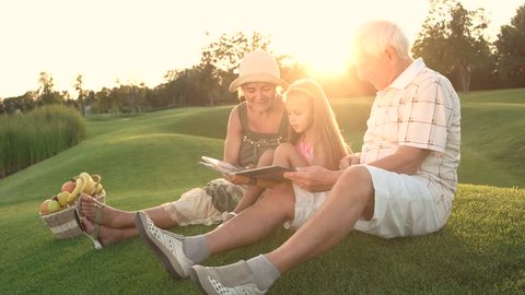 Girl with grandparents outdoors, photo album. People sitting on grass and looking at photos in photo book, basket with fruits. Family on picnic.