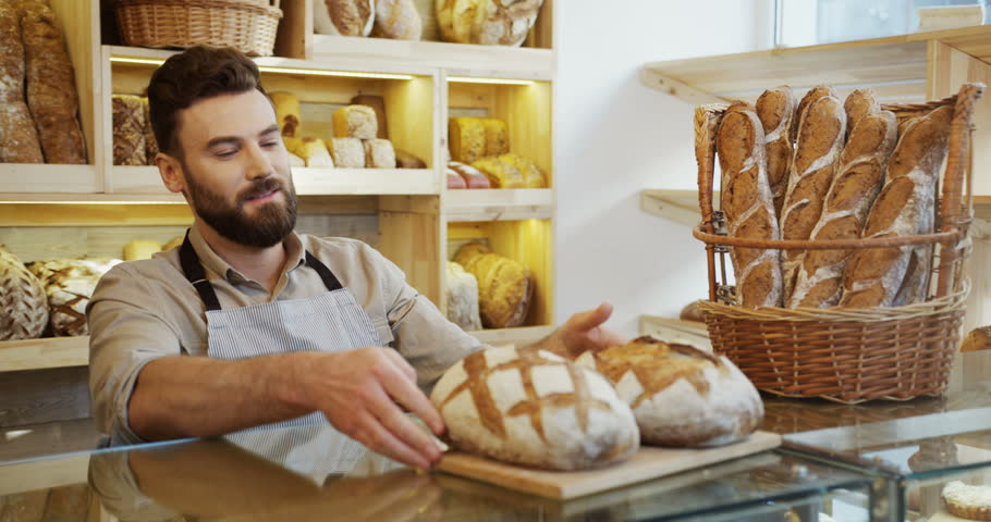 Cucasian attractive young male baker in the shop bringing just baked bread to the counter.