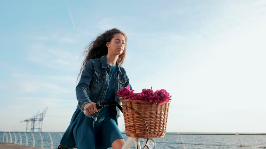 Young attractive woman with curly hair riding vintage bicycle near sea during sunrise or sunset. Girl with flowers in her basket. Romantic scene. Traveler lady. Slow motion.