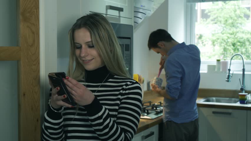 Woman cheating and texting in kitchen partner unaware. Handheld of young girlfriend secretly texting another man whilst boyfriend cooks in background.