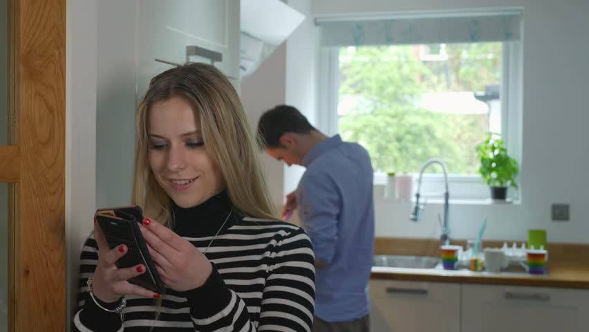 Girlfriend cheating texting slow motion online. Handheld of young girlfriend secretly texting another man whilst boyfriend cooks in background.